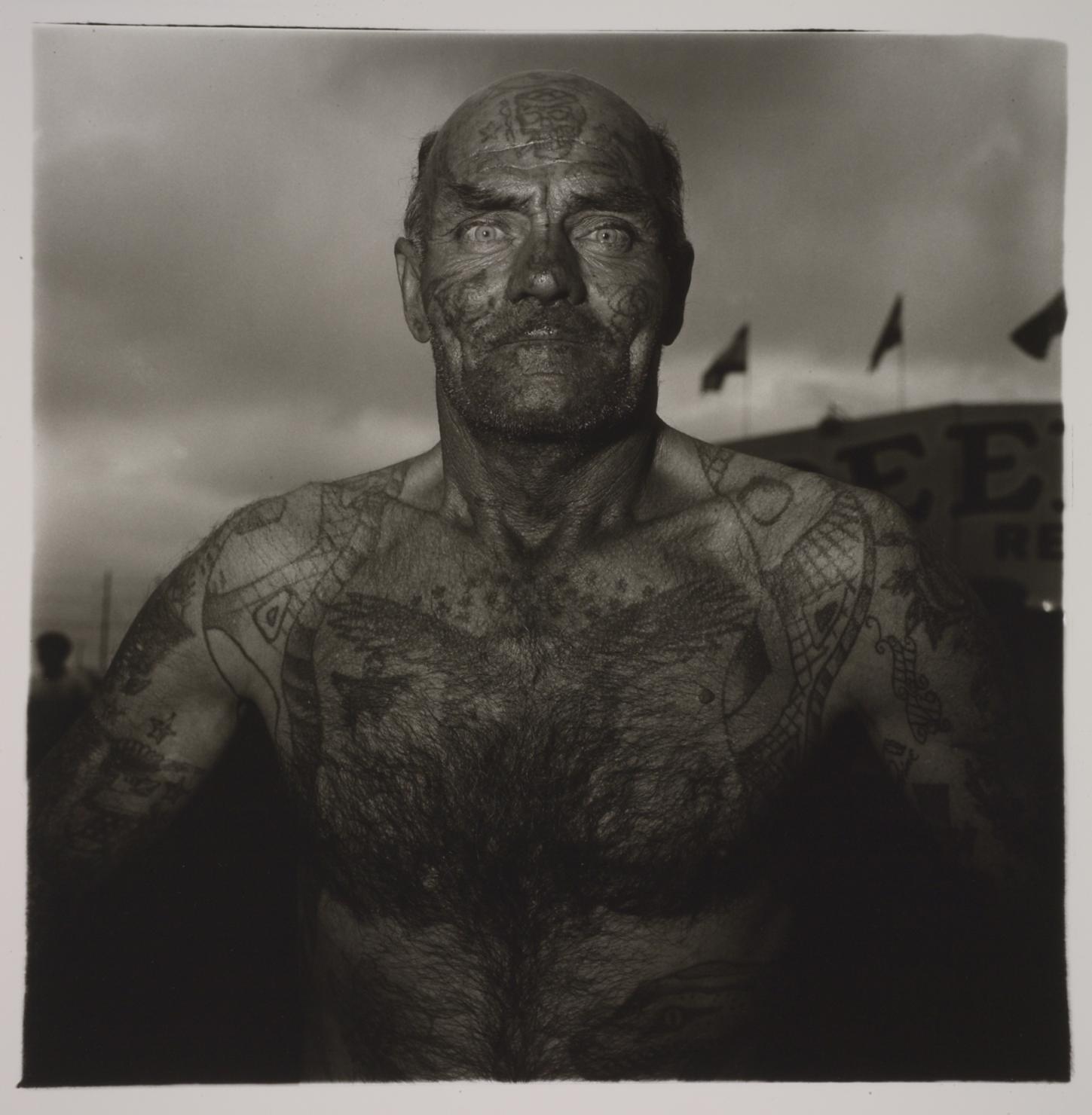 Black and white portrait of a shirtless, bald white man heavily tattooed on his arms, torso, and head 