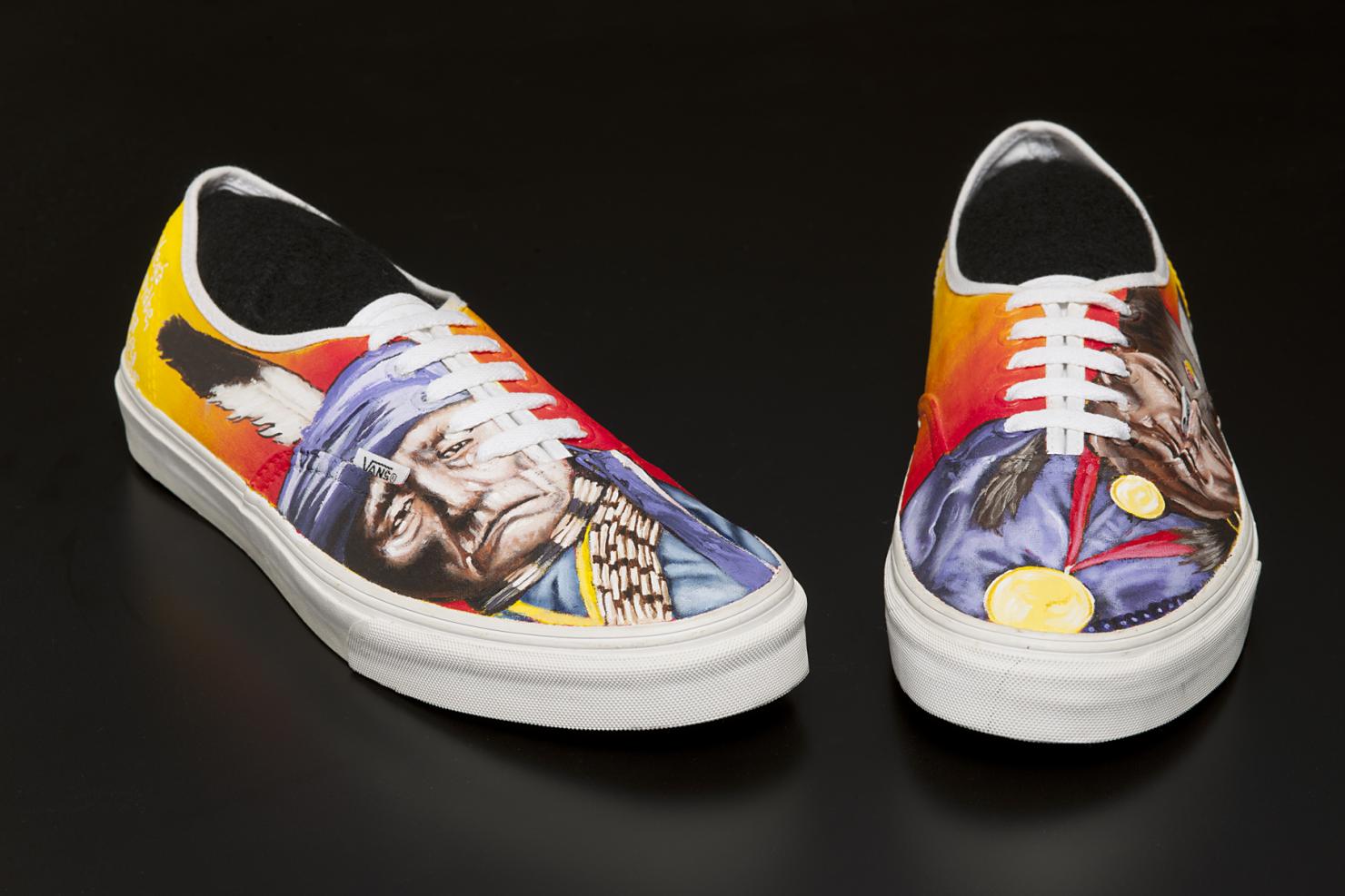 a pair of simple yellow-orange canvas sneakers; the outside of each features a painting of a Native American man with a single feather in his headdress.