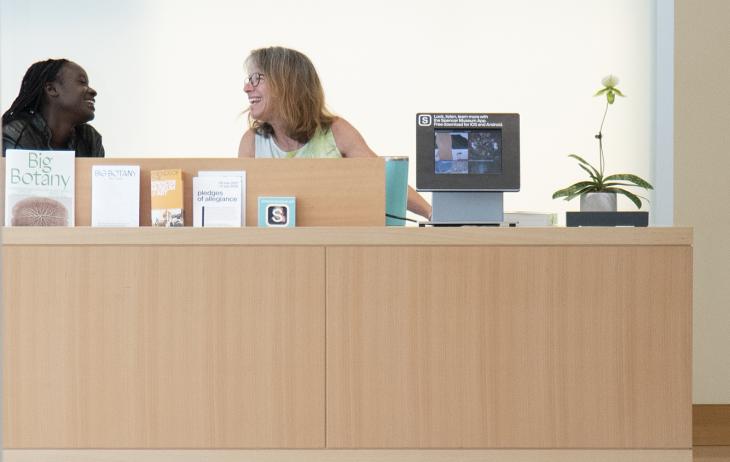Two women sit behind the Museum's Welcome Desk smiling at each other