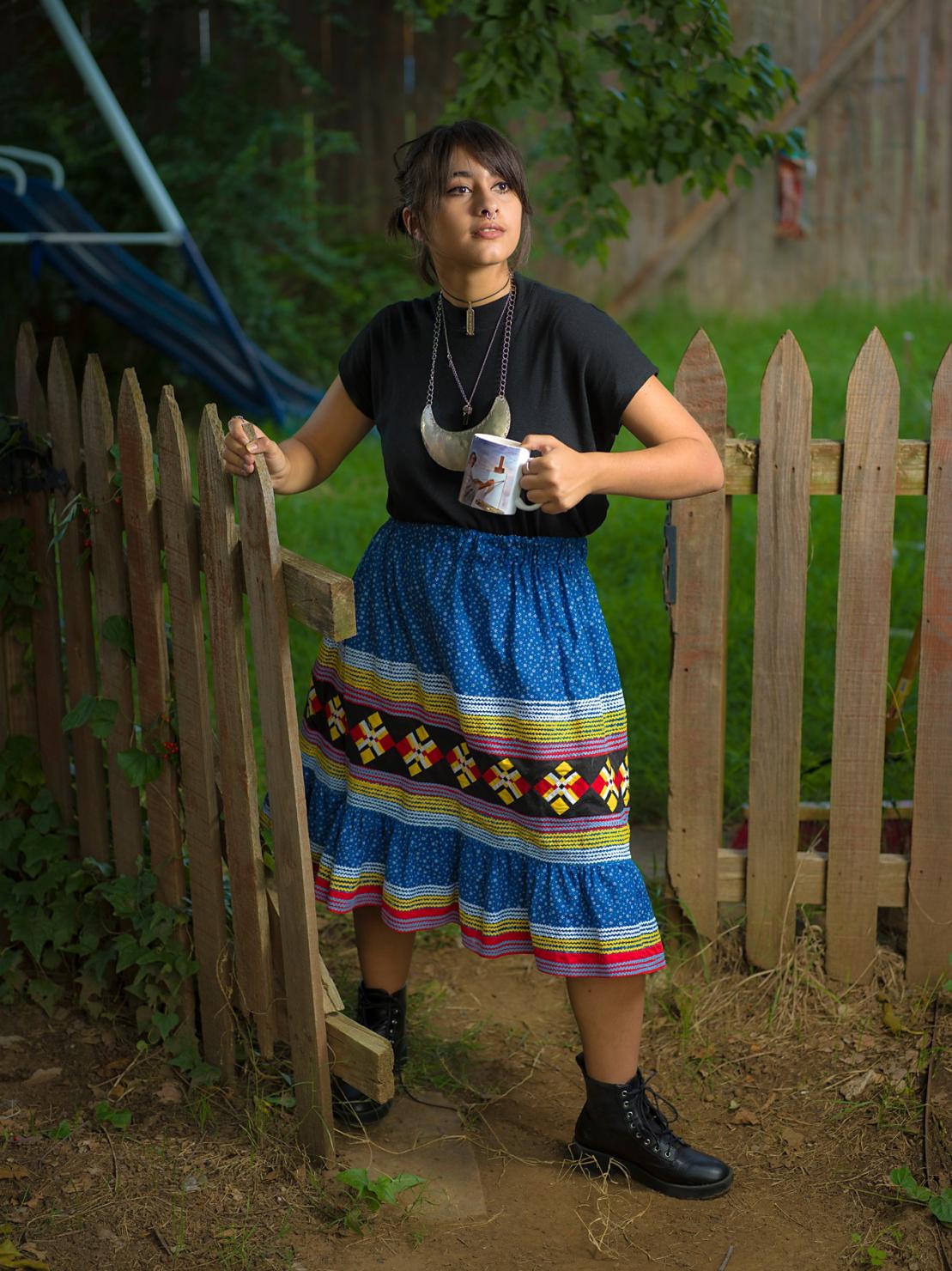 an Indigenous woman carrying a mug in her left hand walks through the gate of a wooden fence; she wears a blue ribbon skirt, black tshirt, and black boots