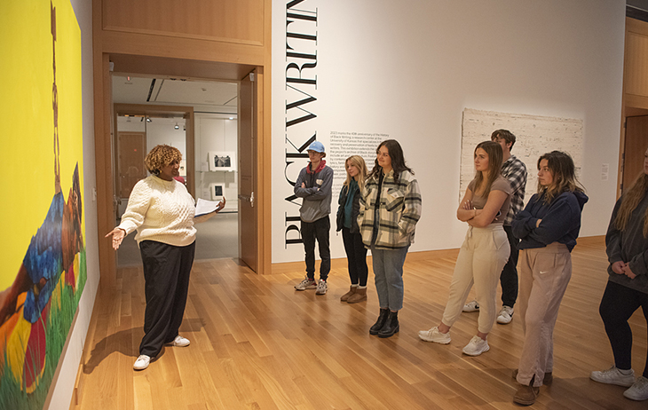 A Black woman with short curly hair talks to a group of students in a gallery while gesturing at a brightly colored painting