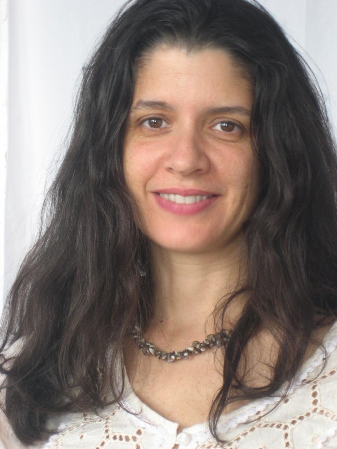 Photographic color portrait of an olive-skinned woman with long dark hair. She is wearing a white blouse and smiling at the camera.