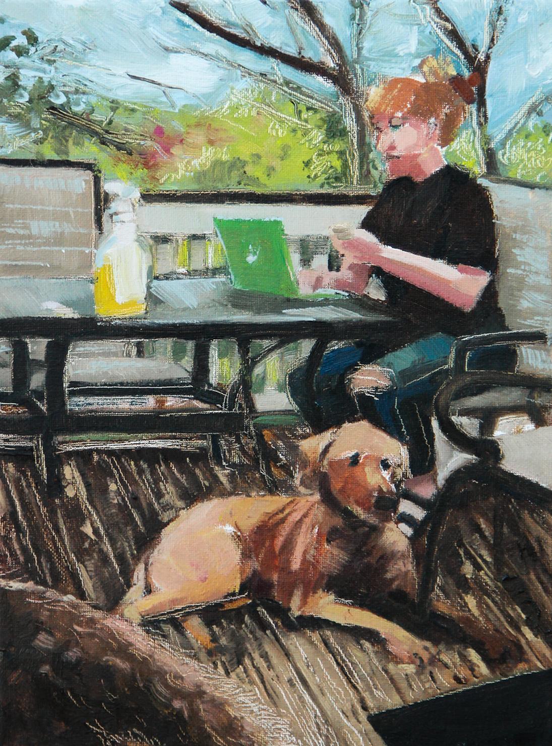 A woman sits outdoors at a table staring at a laptop. A dog sits at her feet.