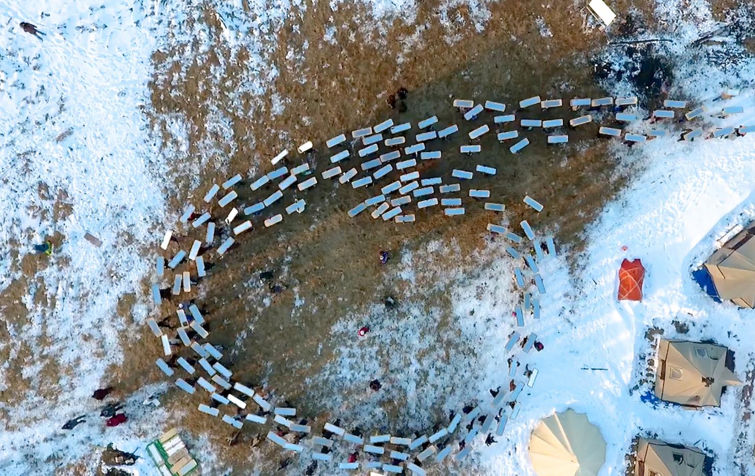 overhead view of a partially snow-covered landscape with a group of people carrying silver rectangles into the shape of a six; a few tents are in the lower right corner