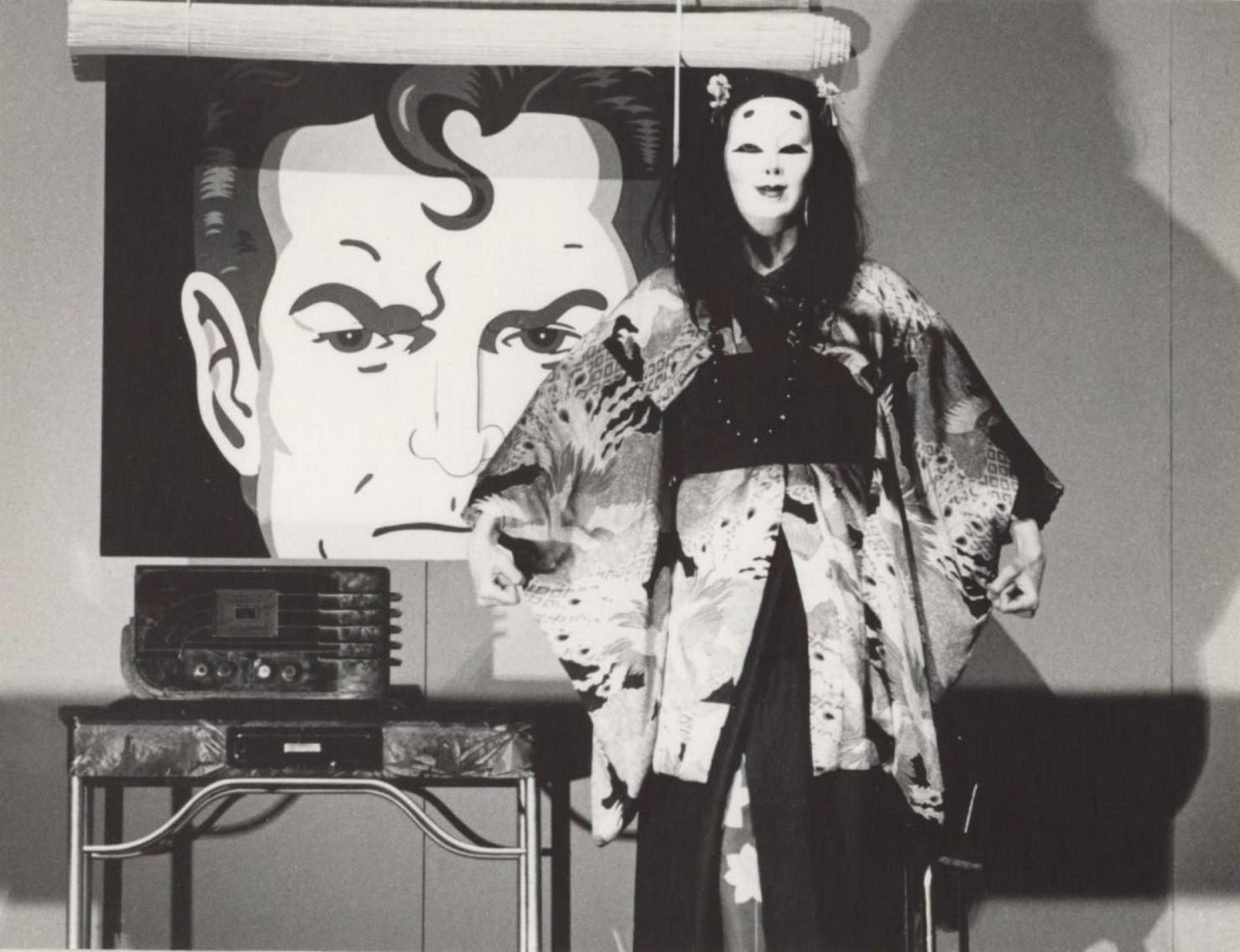 Black and white image with a cartoon depiction of Superman in the background. In the foreground an actor wears a mask and a kimono and listens to an old-fashioned radio.