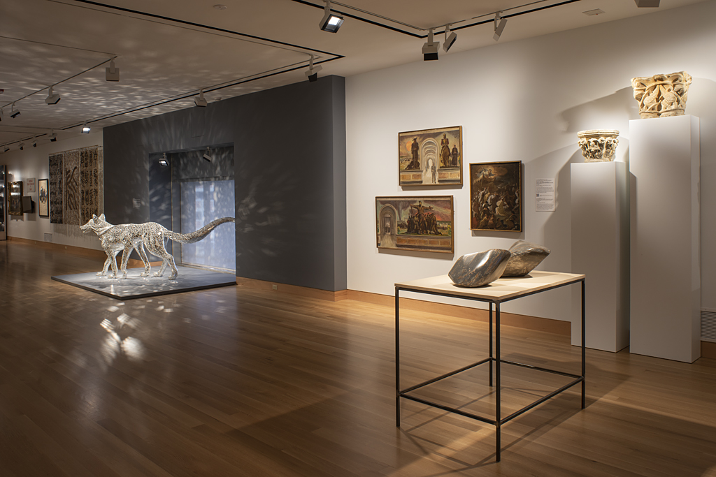 A gallery with paintings on the wall, a pedestal with two irregularly shaped sculptures, and in the background a wolf sculpture