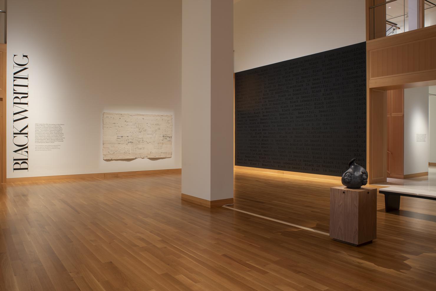 A large gallery with wood floors, one all black wall, the words 'Black Writing' in large text on the other wall
