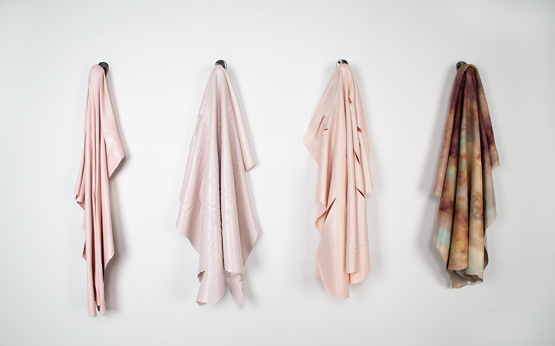 Four textiles hang on equally spaced hooks on a white wall. The first three textiles are shades of pink and the final one is various shades of brown.