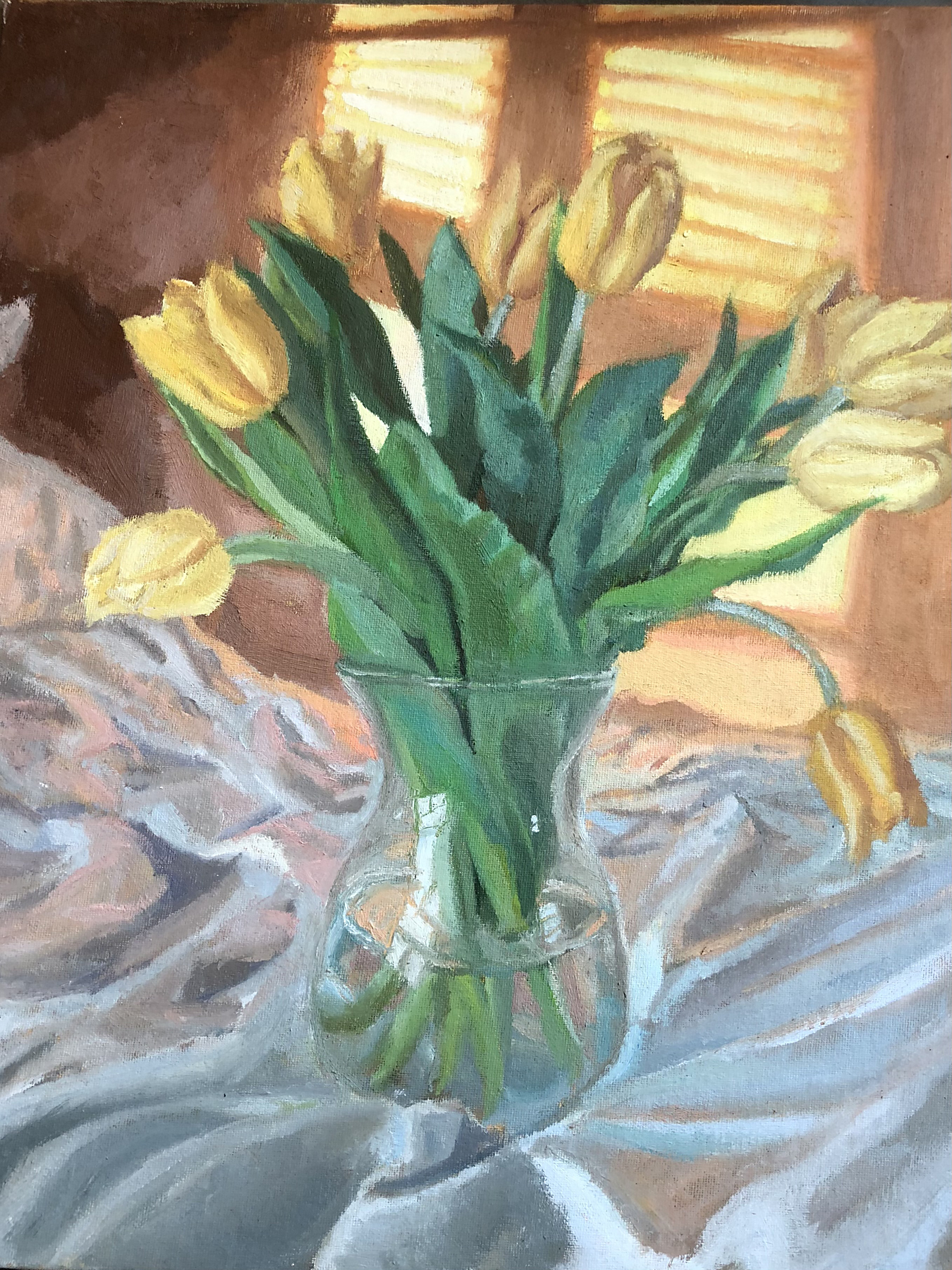 A vase of yellow tulips sits on a white tablecloth with light streaming in the window behind it