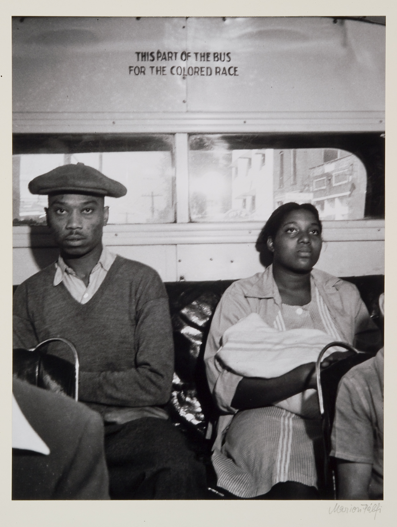 Black-and-white image of an African-American male and female seated on a city bus. Above their heads is printed, “THIS PART OF THE BUS // FOR THE COLORED RACE.”