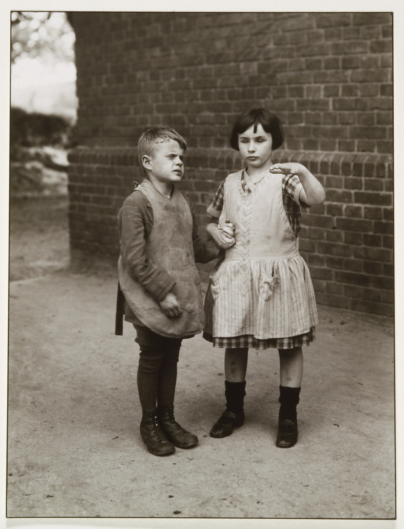 Two children (identified as blind) holding hands. The girl on the right has her proper left arm raised. There is a brick wall behind the two children.
