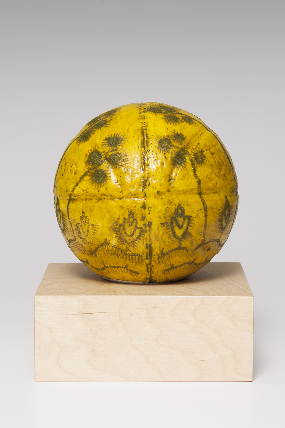 A ceramic sculpture in the shape of a basketball and covered in encaustic. The outlines of the basketball are visible, as well as the logo "Wilson" on the sides. The color is dark yellow and the surface is decorated with lines made up of small circles pressed into the surface, then rubbed with greenish brown pigment. Near the bottom are rows of wavy lines. Extending from the top wavy line is a row of short, tulip shaped flowers alternating with very long stems that reach to the top third of the ball and are