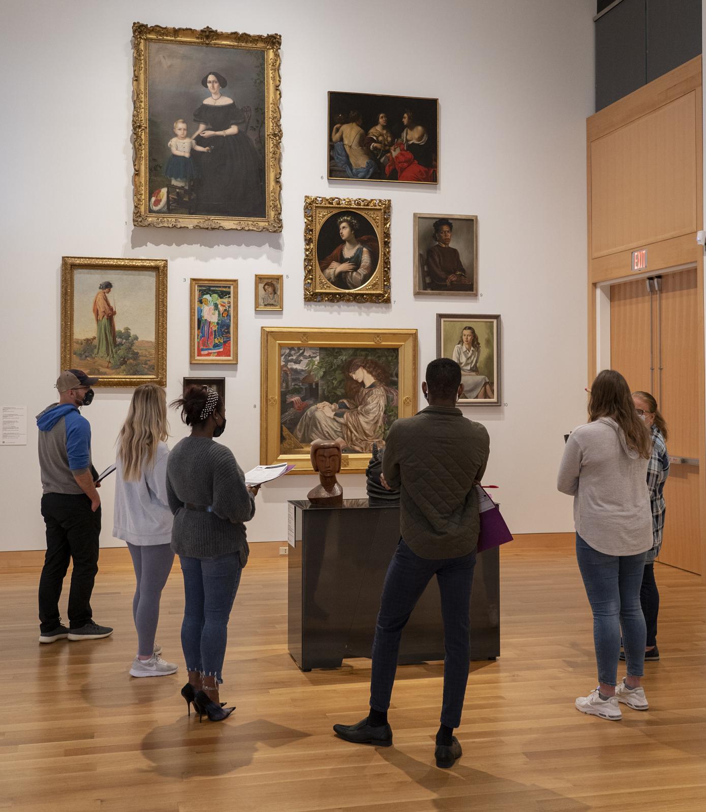 A group of visitors stands in a semicircle looking at a wall of artwork.