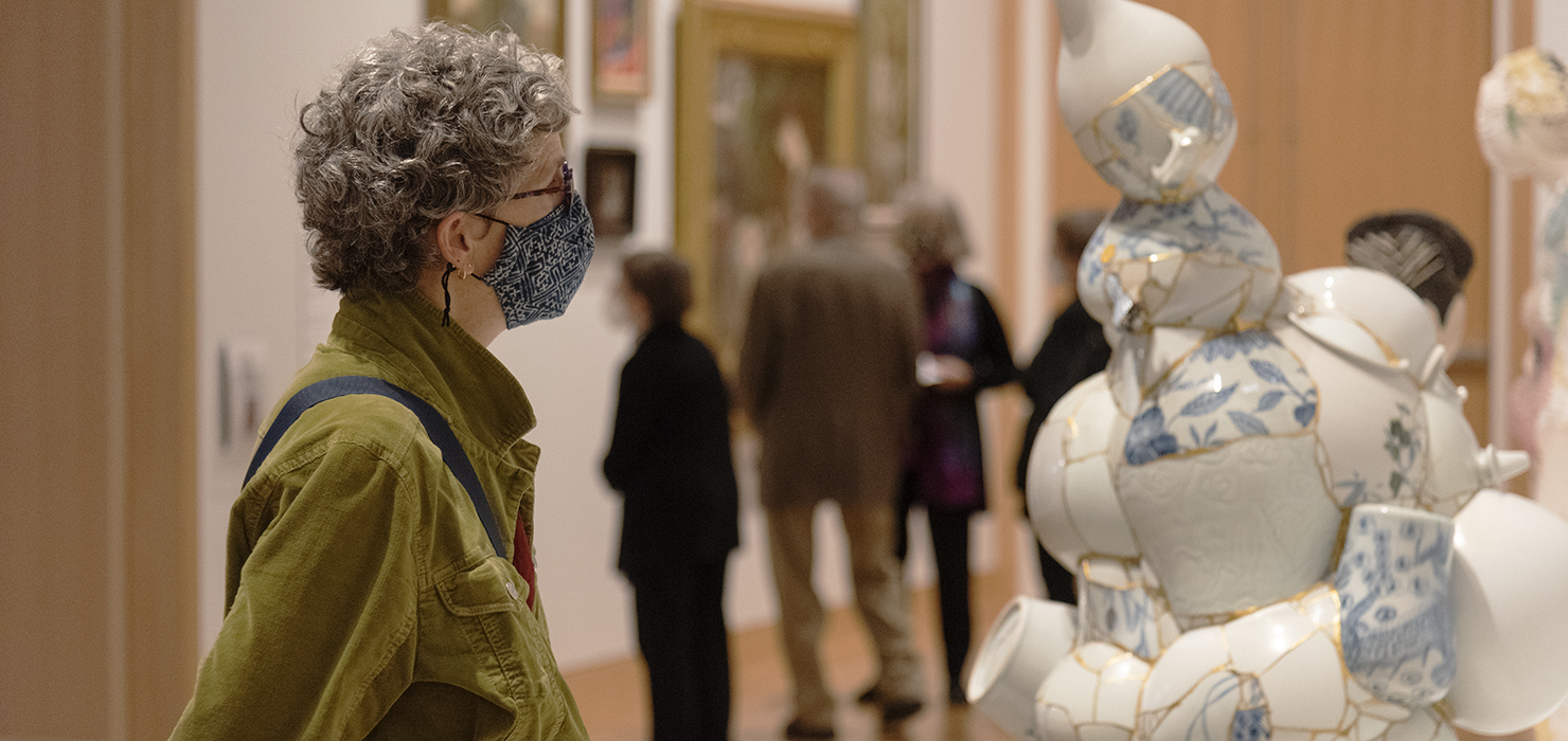 A person with short curly hair wearing glasses and a face mask stares at a ceramic sculpture that is white, gold, and blue.