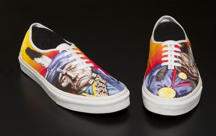 a pair of simple yellow-orange canvas sneakers; the outside of each features a painting of a Native American man with a single feather in his headdress.