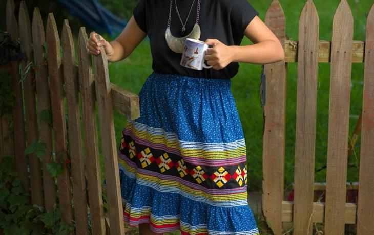 an Indigenous woman carrying a mug in her left hand walks through the gate of a wooden fence; she wears a blue ribbon skirt, black tshirt, and black boots