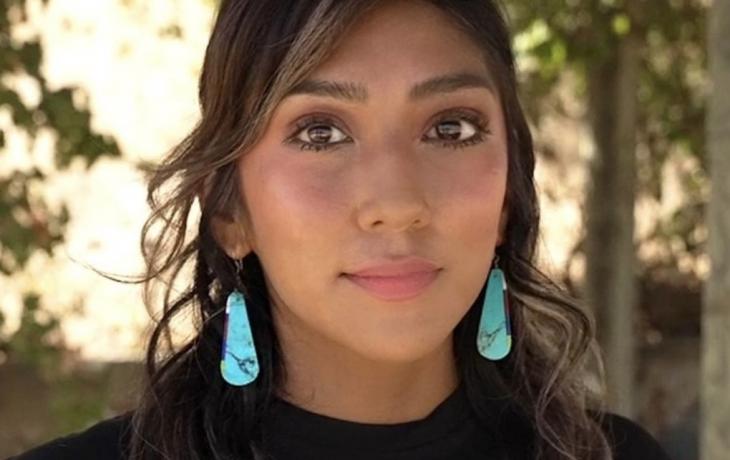 Headshot of an Indigenous woman with long, wavy brown hair wearing large pear-shaped turquoise earrings and a black and turquoise tshirt