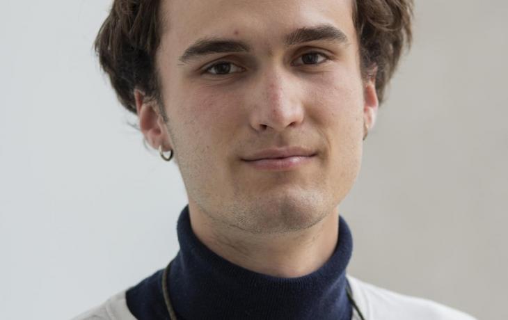 Color photograph of a young person with short brown hair and small hoop earrings identified as Spencer Reeve wearing a white shirt over a dark blue turtleneck