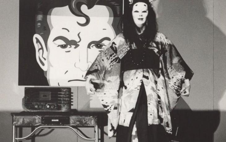 Black and white image with a cartoon depiction of Superman in the background. In the foreground an actor wears a mask and a kimono and listens to an old-fashioned radio.