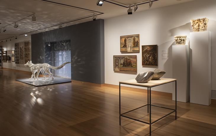 A gallery with paintings on the wall, a pedestal with two irregularly shaped sculptures, and in the background a wolf sculpture