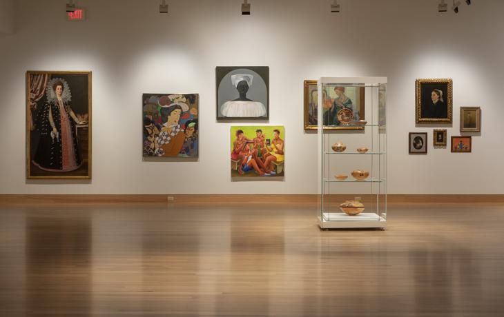 A vertical glass case holds pottery in front of a white wall with many paintings