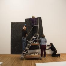 Artist Sandy Winters, Assistant Collections Manager Sarah Schroeder, and Head of Collections Sofia Galarza Liu begin to affix a portion of <i>Long Night's Journey into Day</i> in the Perkins Central Court