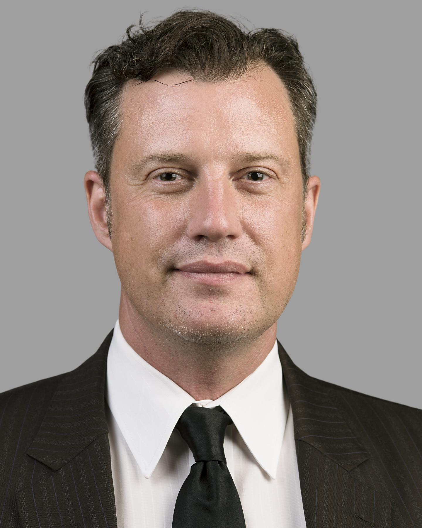 A white man with cropped dark hair wears a black tie and smiles at the camera