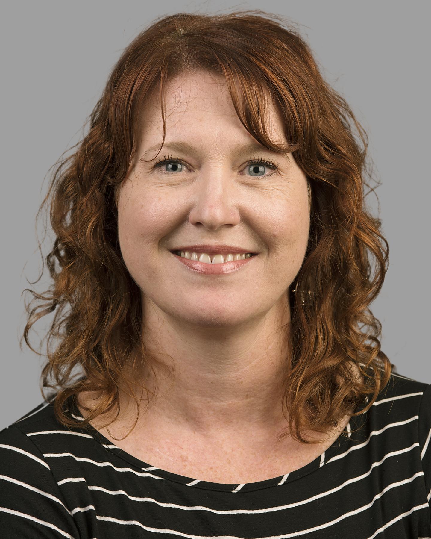 A white woman with red curly hair wears a striped black shirt and smiles at the camera