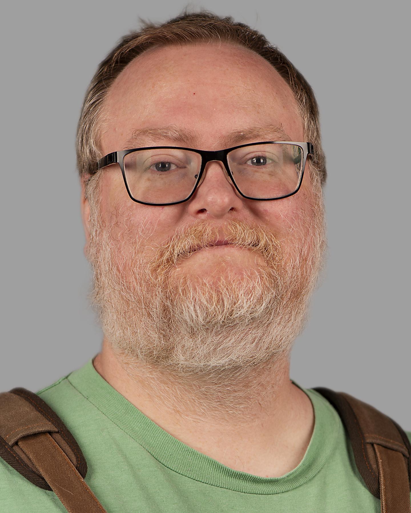 A bearded white man smiles at the camera, he wears black framed glasses and a green shirt