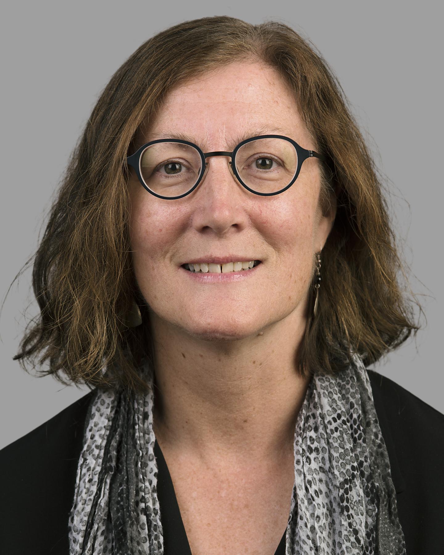 A white woman with short brown hair wears black framed glasses and a scarf with gray and black dots on it, she smiles at the camera