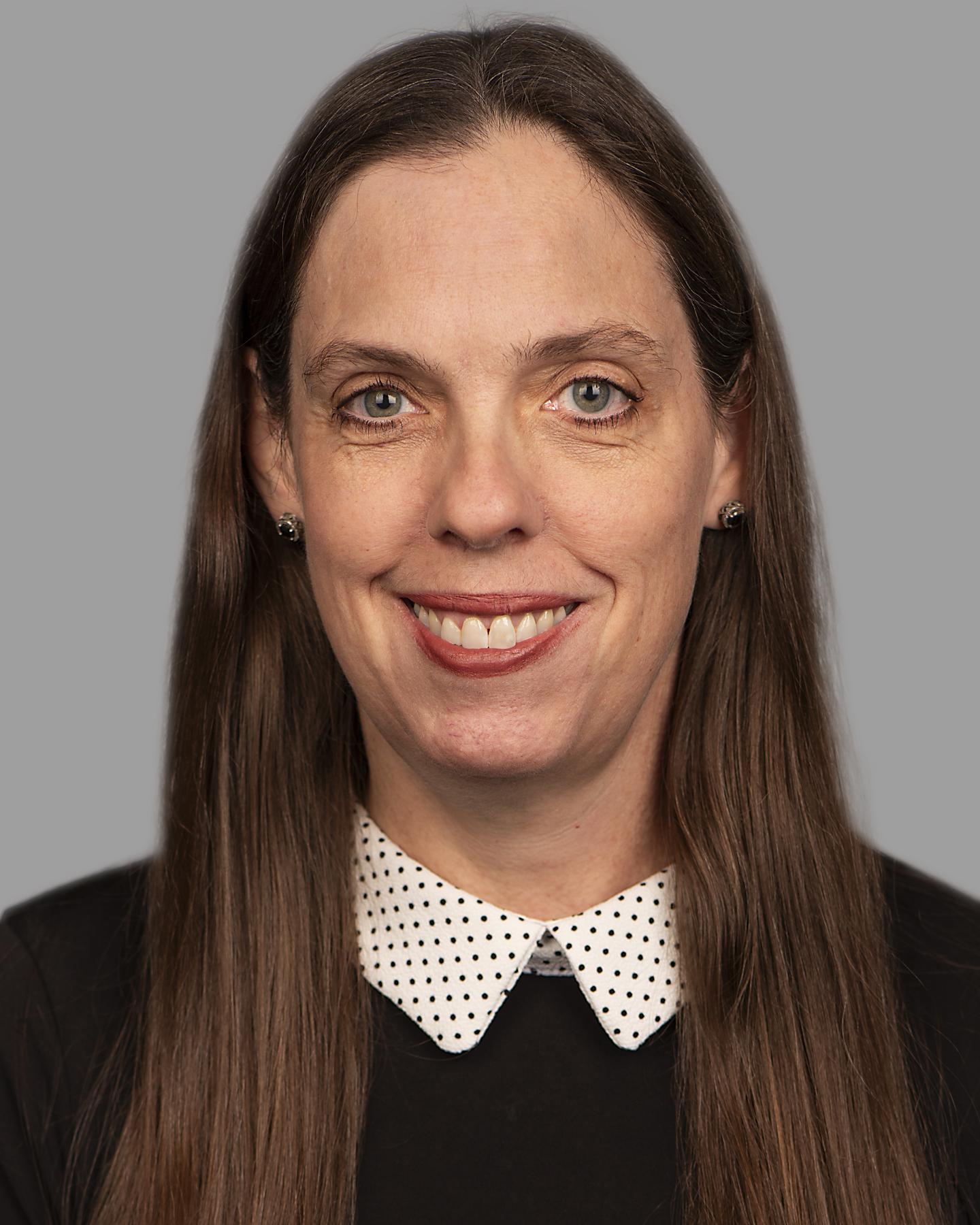 A white woman with long brown hair smiles at the camera, she wears a black shirt with a white and black polka dot collar