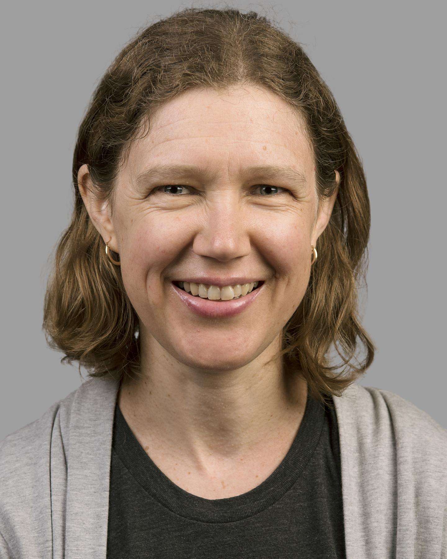 A white woman with shoulder-length brown hair smiles at the camera