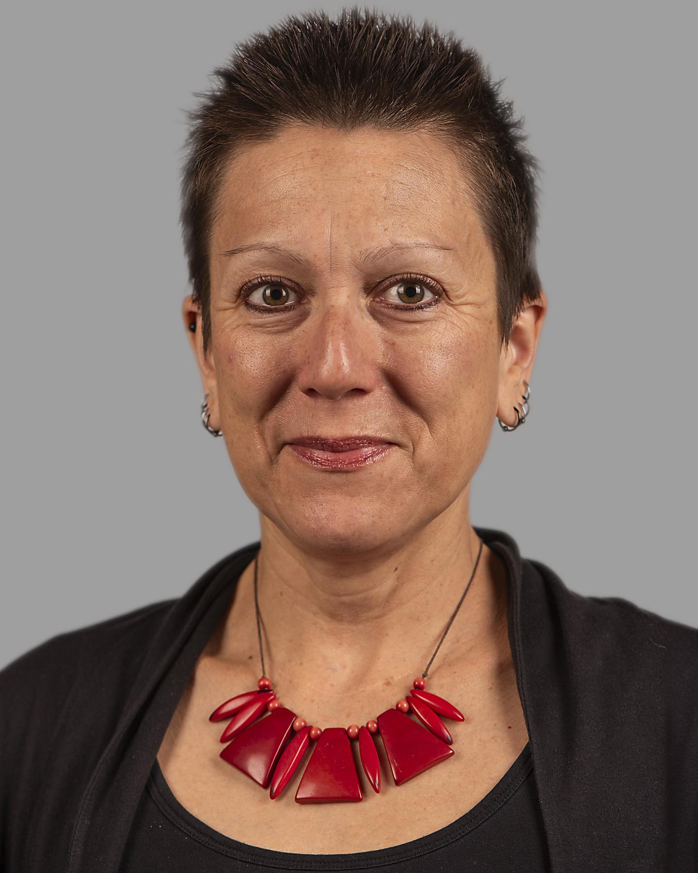 A white woman with short cropped brown hair smiles at the camera, she wears four hoop earrings on each ear and a large red geometric necklace