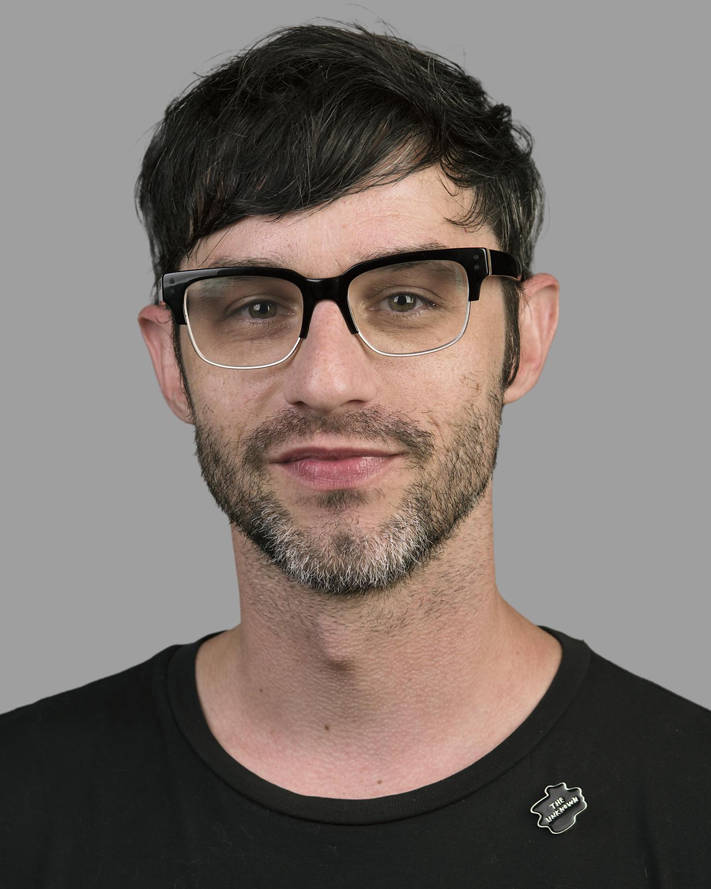 A white bearded man with cropped black hair wears black square framed glasses and smiles at the camera