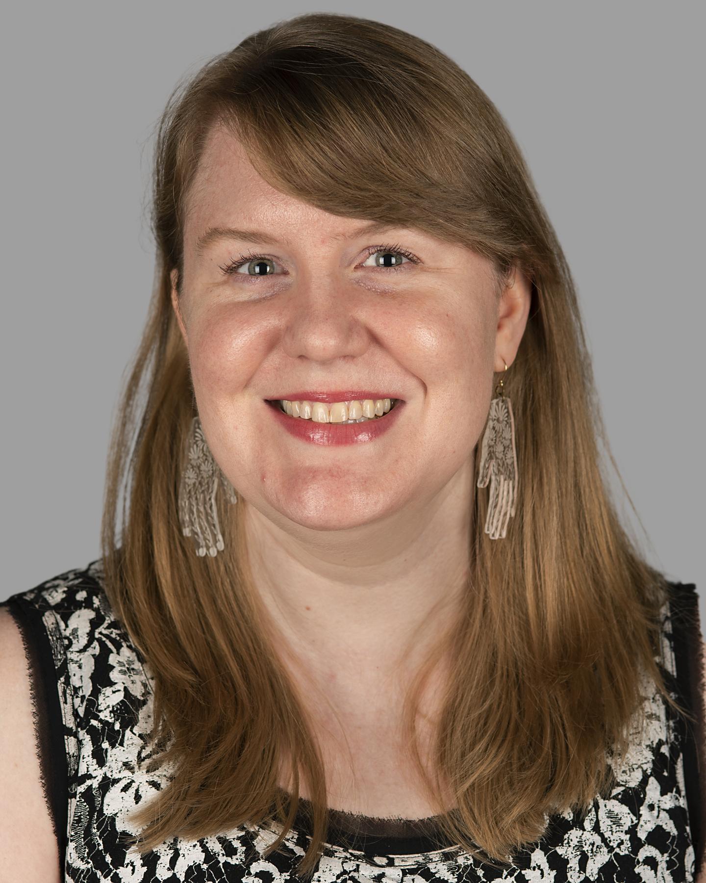 A white woman with medium-length blonde hair wears earrings shaped like hands and dark pink lipstick; she smiles at the camera