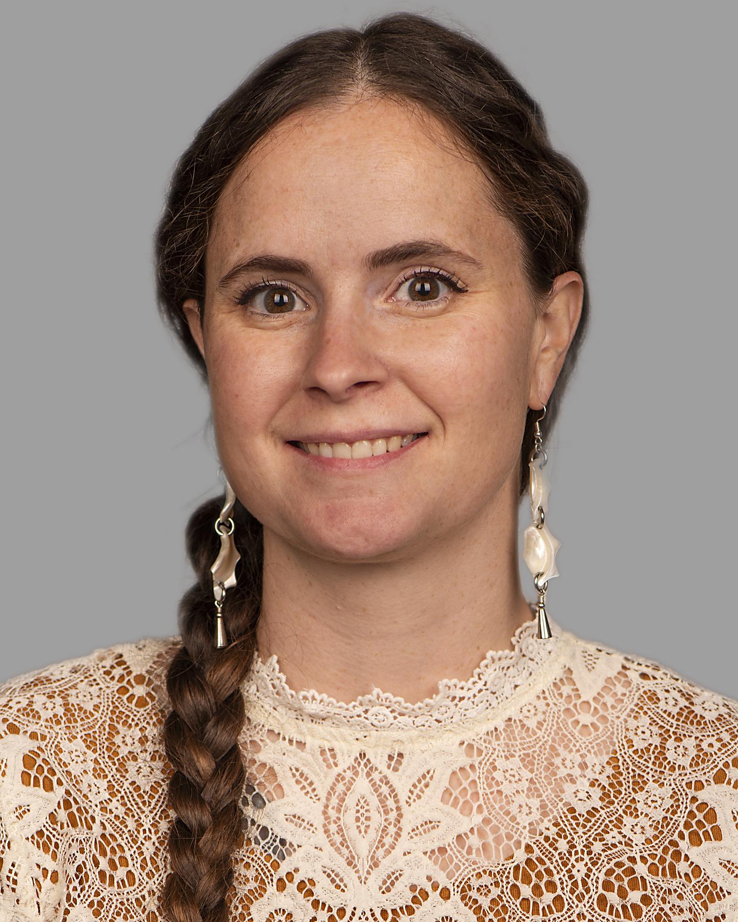 A light-skinned Indigenous woman with brown hair in a long braid over her right shoulder wears long shell earrings and a lace top; she smiles at the camera 