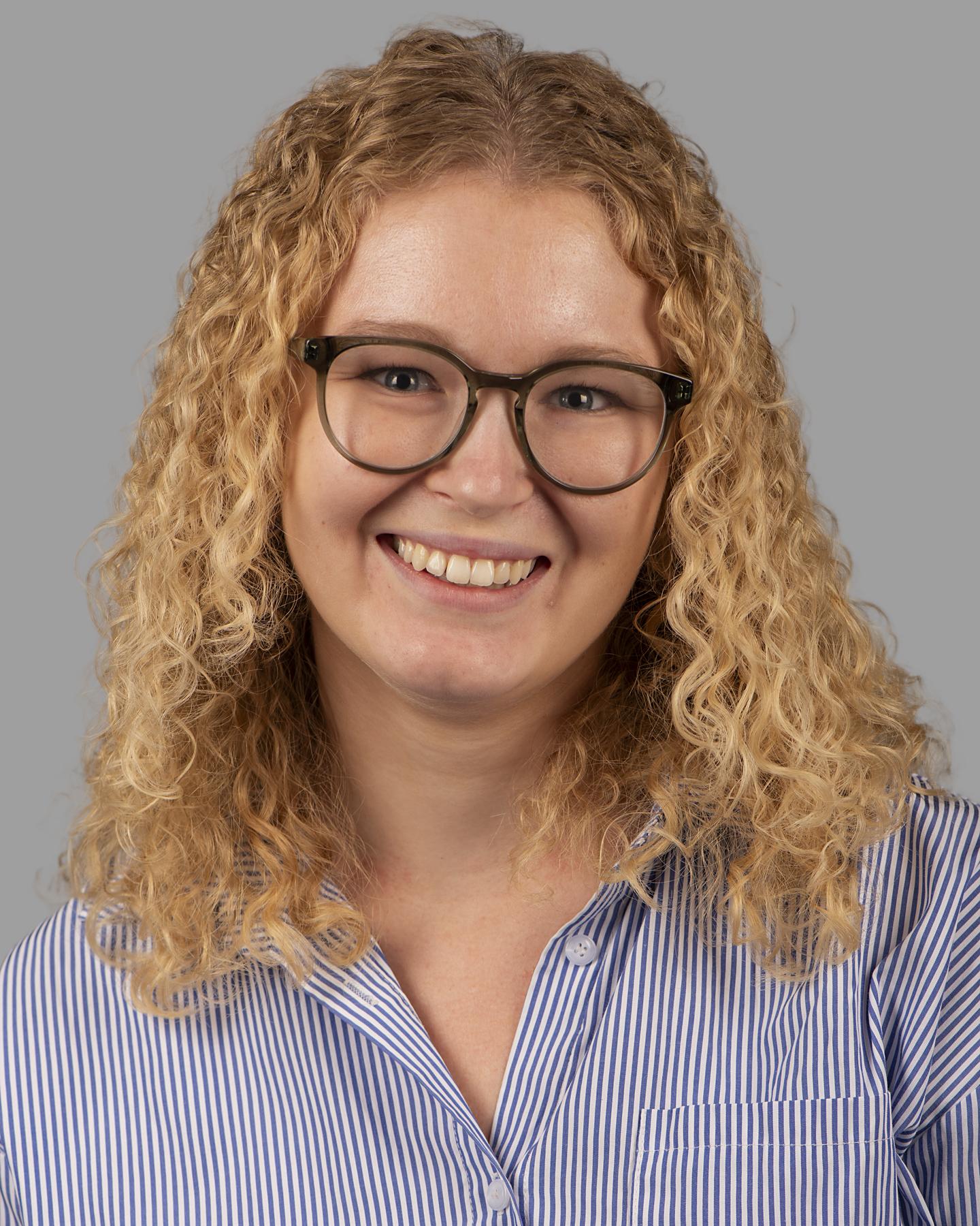  A white woman with shoulder length curly blonde hair wears circular framed glasses and smiles at the camera