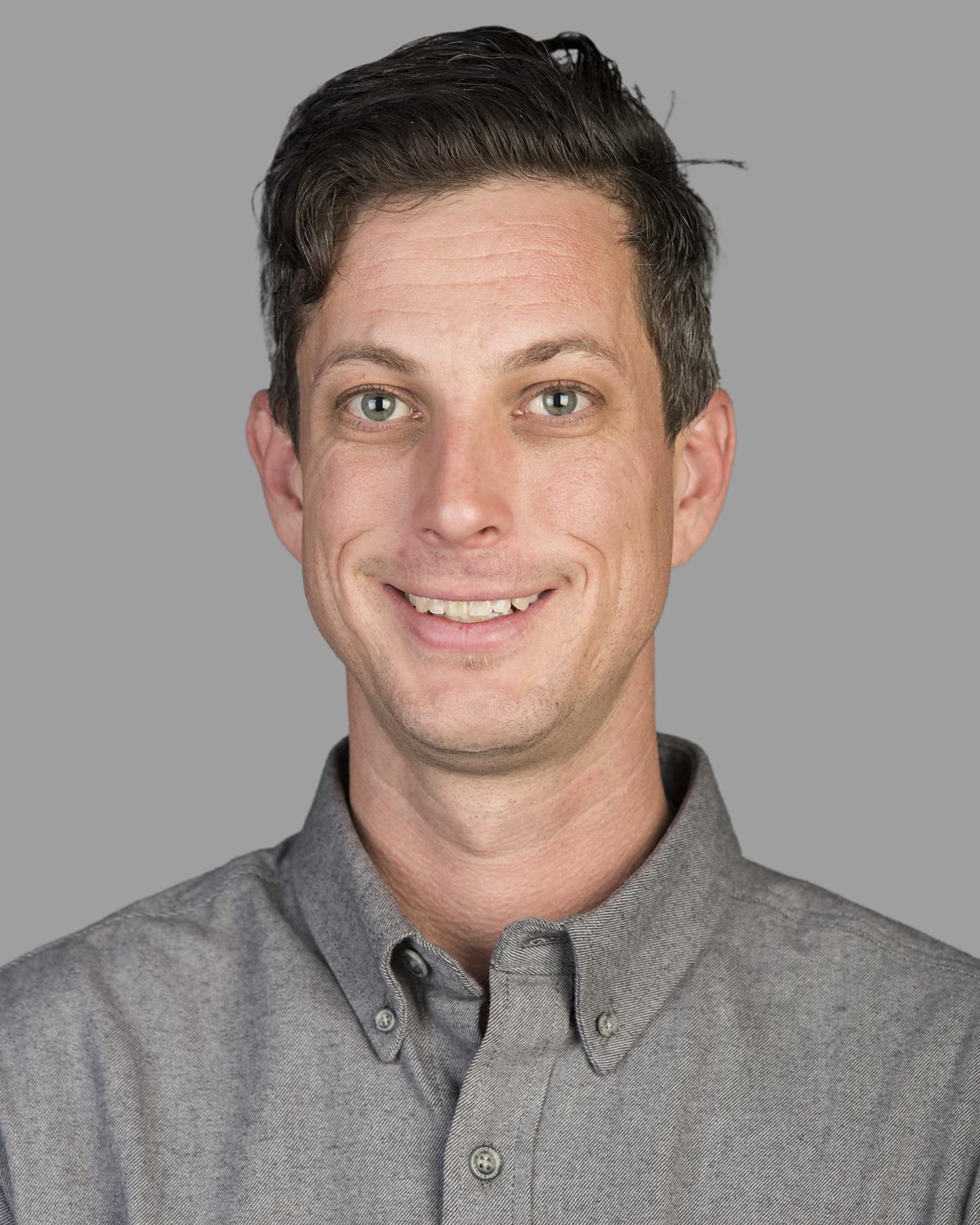 A white man with cropped black hair wears a gray collared shirt and smiles at the camera
