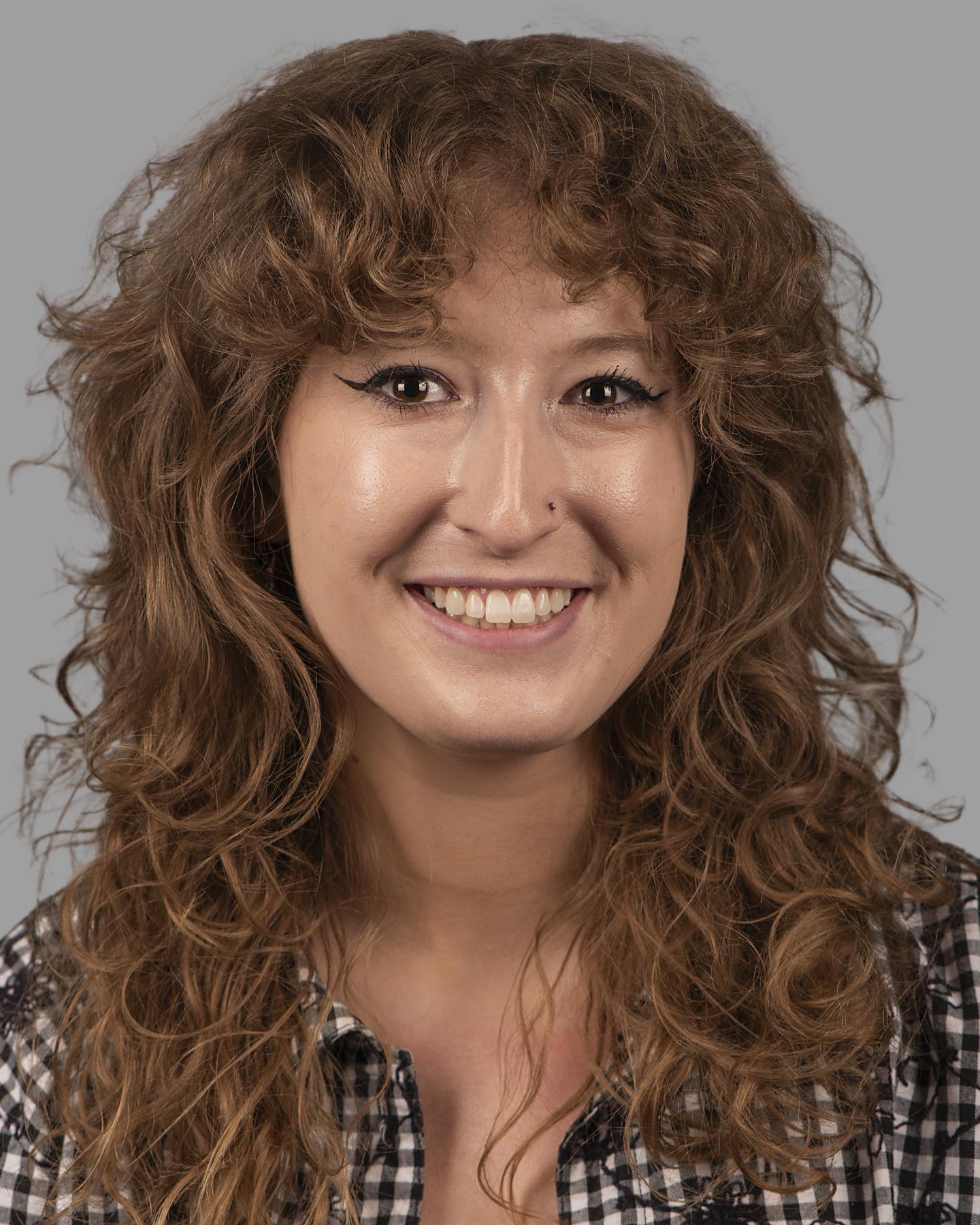 A white woman with long curly brown hair wears a black gingham shirt and smiles at the camera