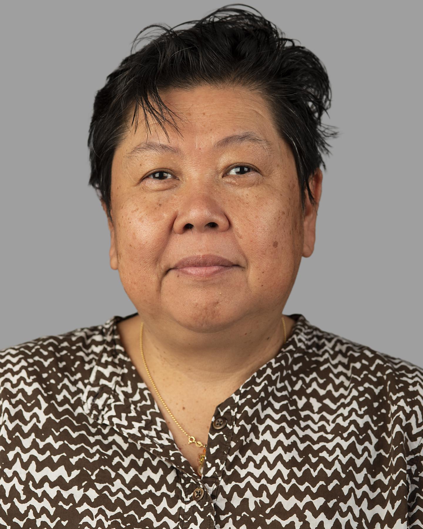 An Asian woman with cropped black hair wears a white shirt with a brown zig zag pattern on it; she smiles at the camera