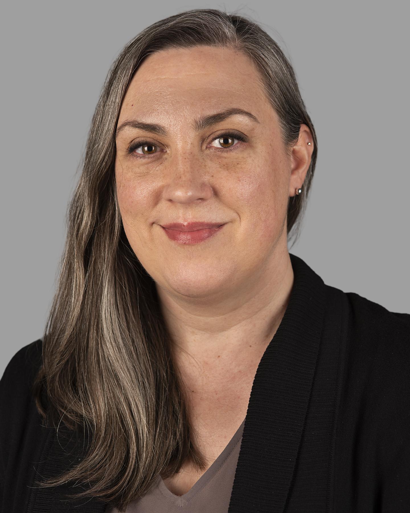 A white woman has long brown hair with streaks of gray throughout, she wears shiny light red lipstick and smiles as the camera