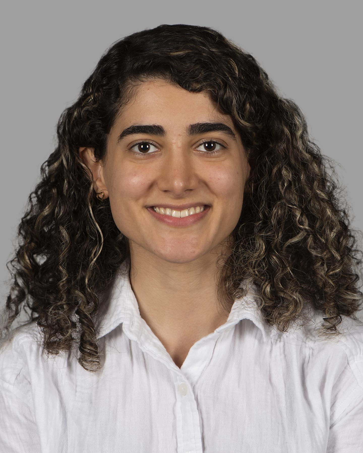 A light brown skinned woman with shoulder-length dark curly hair wears a white button up shirt and smiles at the camera