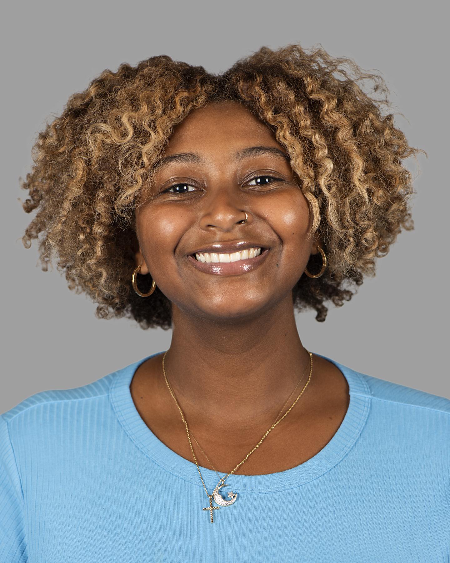 A Black woman has short light brown coily hair and wears a bright blue shirt; she smiles at the camera