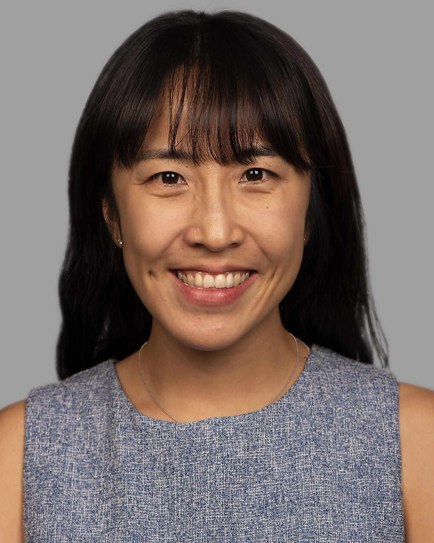 An Asian woman with long black hair wears a sleeveless tweed top and smiles at the camera