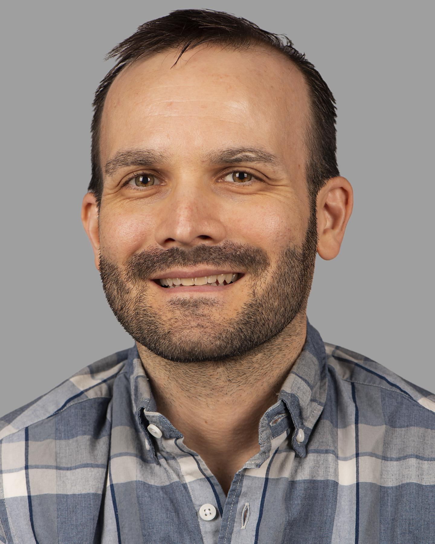 A white man with short black hair and facial hair wearing a plaid collared shirt smiles at the camera