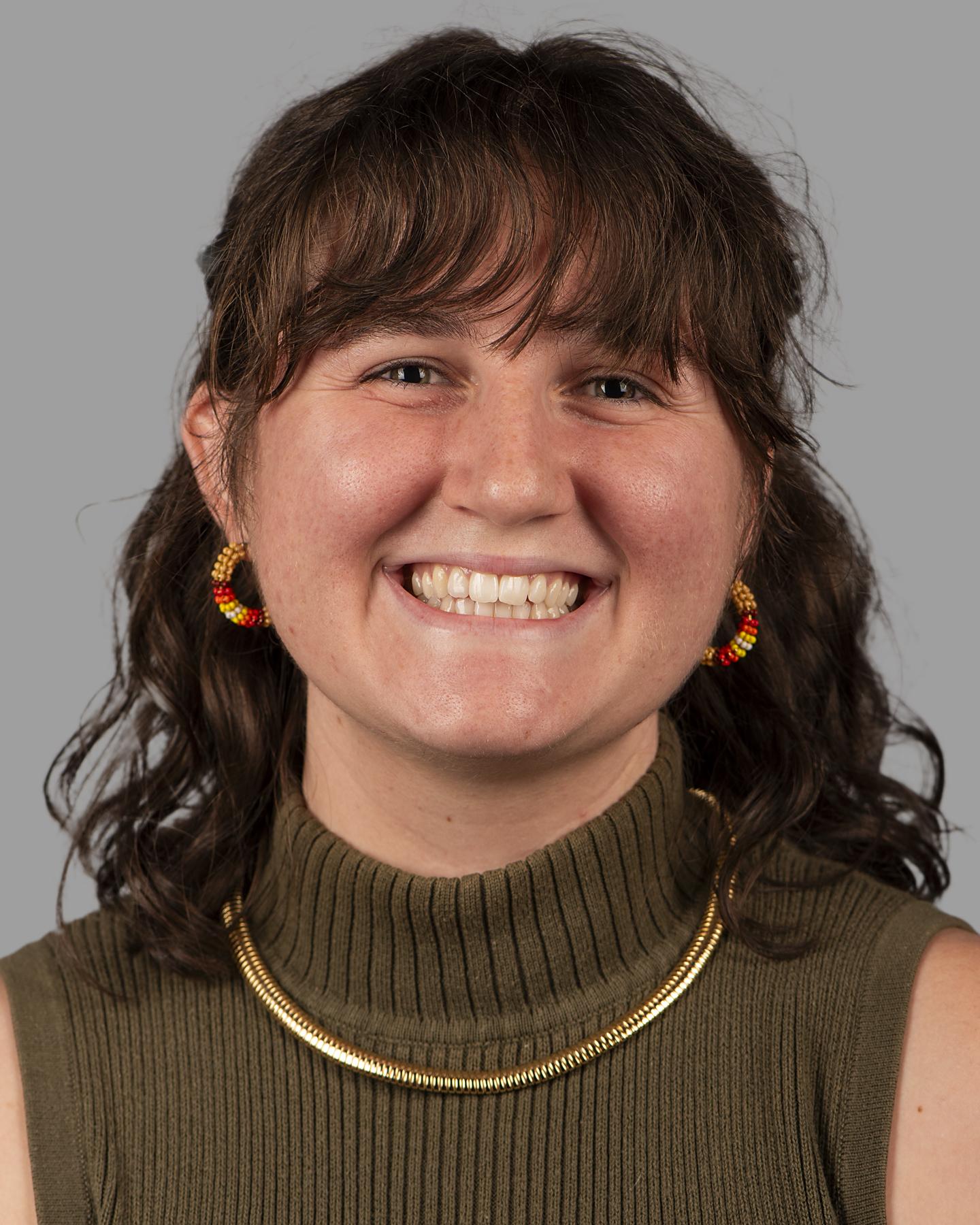 A white Indigenous woman with shoulder-length brown curly hair wears brown and red beaded hoop earrings and smiles at the camera 