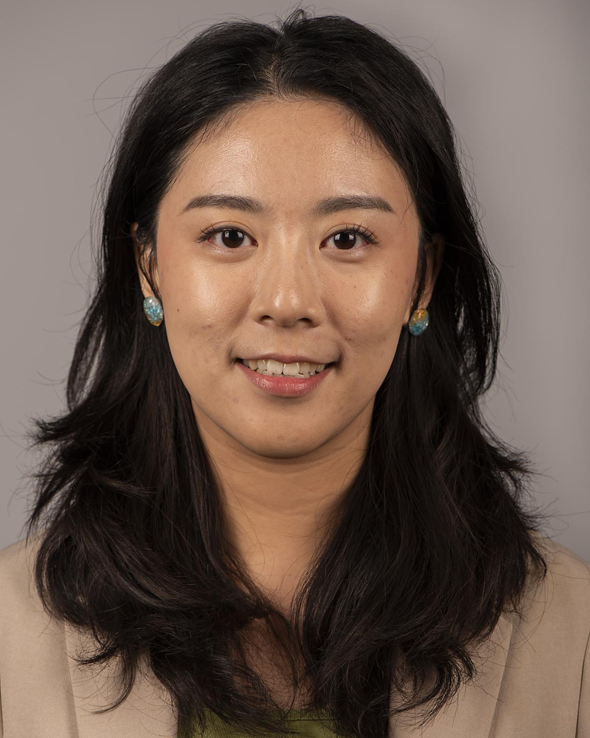 An Asian woman with medium-length black hair wears a tan blazer and smiles at the camera