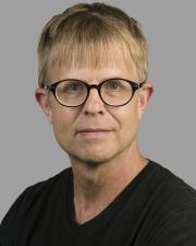 A white man with short dirty blonde hair wears black framed circular glasses and smiles at the camera