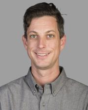 A white man with cropped black hair wears a gray collared shirt and smiles at the camera 