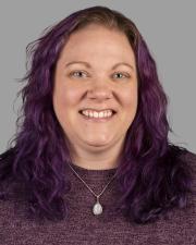 A white woman with medium-length wavy purple hair wears an opal necklace and smiles at the camera